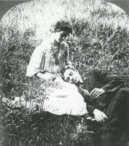 a young couple recline in the grass. the young man's head rests in his female companion's lap, while her hand rests in his hair and she gazes down at him.