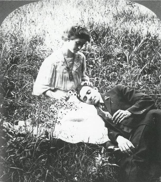 a young couple recline in the grass. the young man's head rests in his female companion's lap, while her hand rests in his hair and she gazes down at him.